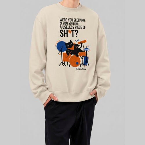 Front view of a male model wearing the Black Scowl beige crewneck sweatshirt. The urban streetwear design features a sarcastic black cat playing drums with an edgy, ironic quote.