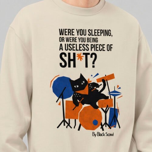Close-up view of the Black Scowl graphic apparel on a cream sweatshirt. The edgy streetwear print displays an existential black cat drummer and dark humor typography.
