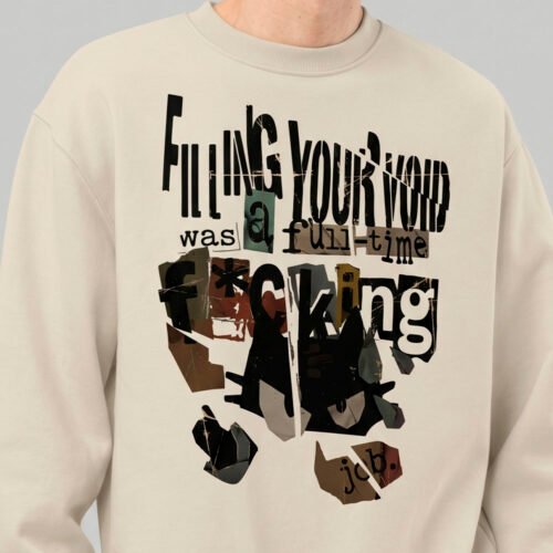 Close-up detail of the Black Scowl beige sweatshirt, showcasing the modern graphic wear print with a dark cat and ransom-note style lettering.