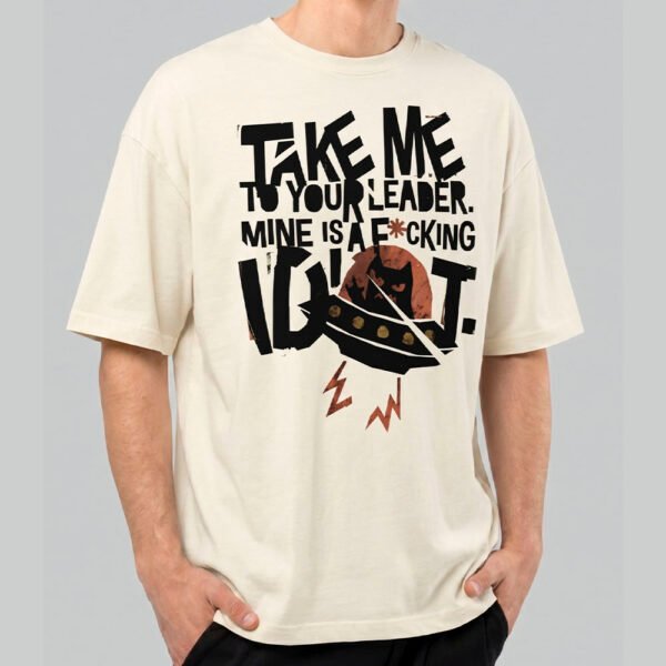 Front view of a male model wearing a Black Scowl cream heavyweight t-shirt featuring a cynical alien cat in a UFO with "Take Me To Your Leader" graphic text.
