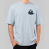 Front view of a male model wearing a Black Scowl light blue oversized t-shirt featuring a small graphic of a cynical black cat surfing on a wave.