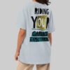 A female model showcasing the back of a Black Scowl light blue streetwear t-shirt with a large "Garbage Expectations" graphic and a relaxed, oversized fit.