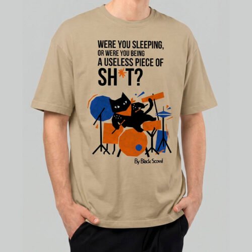 Front view of a male model wearing a Black Scowl sand oversized t-shirt featuring a sarcastic "Were you sleeping or being a useless piece of sh*t?" graphic with a cat drummer.